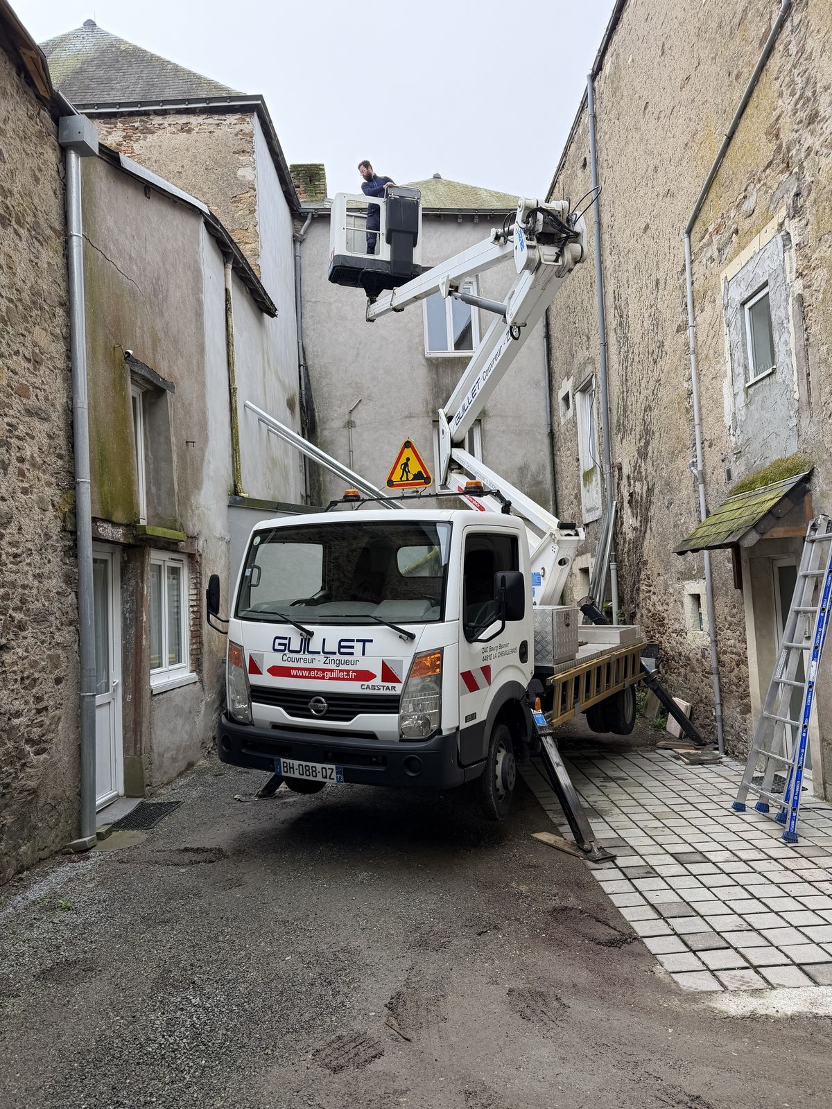 Camion nacelle déployé dans une ruelle étroite, un couvreur en panier au niveau de la toiture